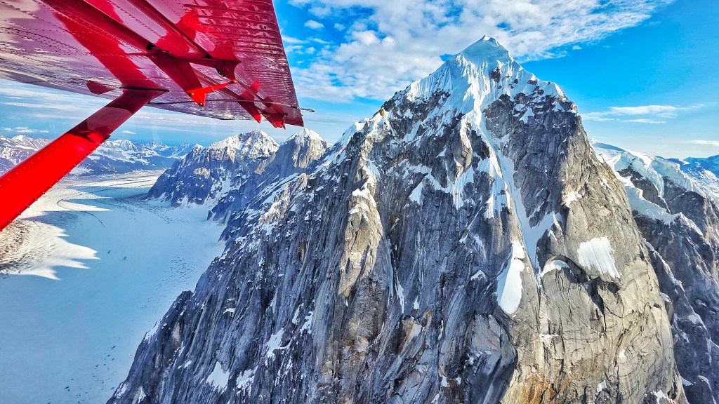 Aerial view of snow-capped mountains with a red airplane wing visible in the foreground against a blue sky.