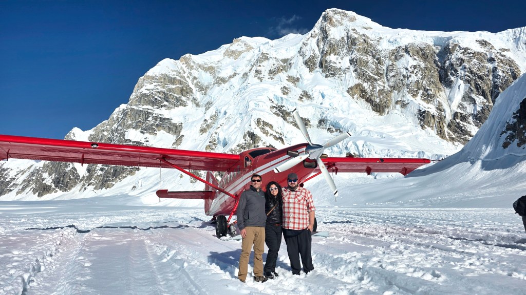 A group of three people standing in the snow next to a red airplane, with towering snow-capped mountains in the background under a clear blue sky.